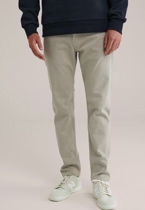 Mand iført lysegrå bukser, hvide sneakers og en mørk langærmet skjorte, der står mod en ensfarvet baggrund.