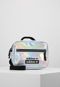 Bolso Adidas plateado iridiscente de forma rectangular, con un bolsillo frontal con cremallera y una correa negra. Incluye dos parches de logo negros.