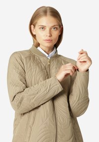 Chaqueta acolchada beige con cuello alto, patrón floral texturizado y cremallera frontal. Presenta mangas largas con puños de canalé.
