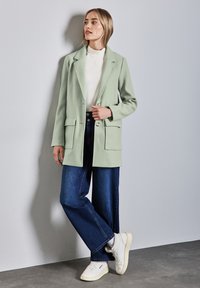 Blazer verde menta con rever a scalino, due tasche frontali e chiusura con un bottone, abbinato a jeans blu scuro larghi e sneakers bianche.