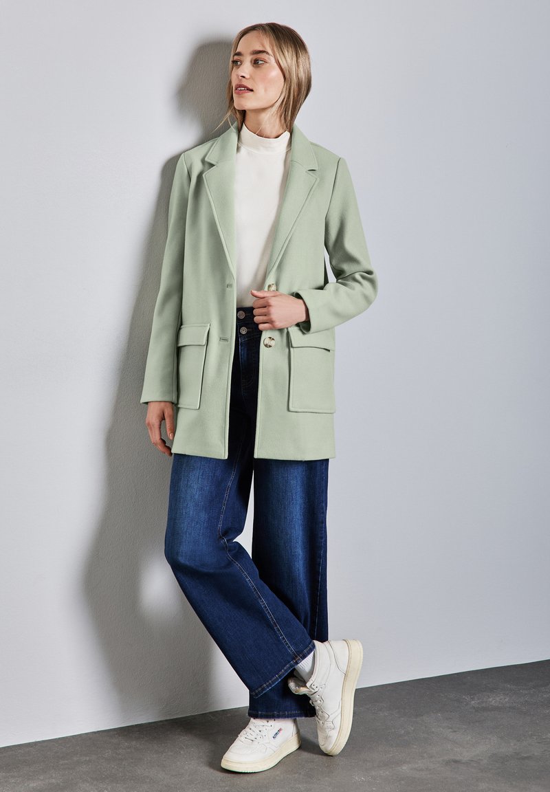 Blazer verde menta con rever a scalino, due tasche frontali e chiusura con un bottone, abbinato a jeans blu scuro larghi e sneakers bianche.