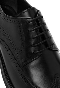 Zapato de vestir negro de cuero con un diseño clásico de brogue, punta redondeada y perforaciones decorativas a lo largo de las costuras y el área de los cordones.