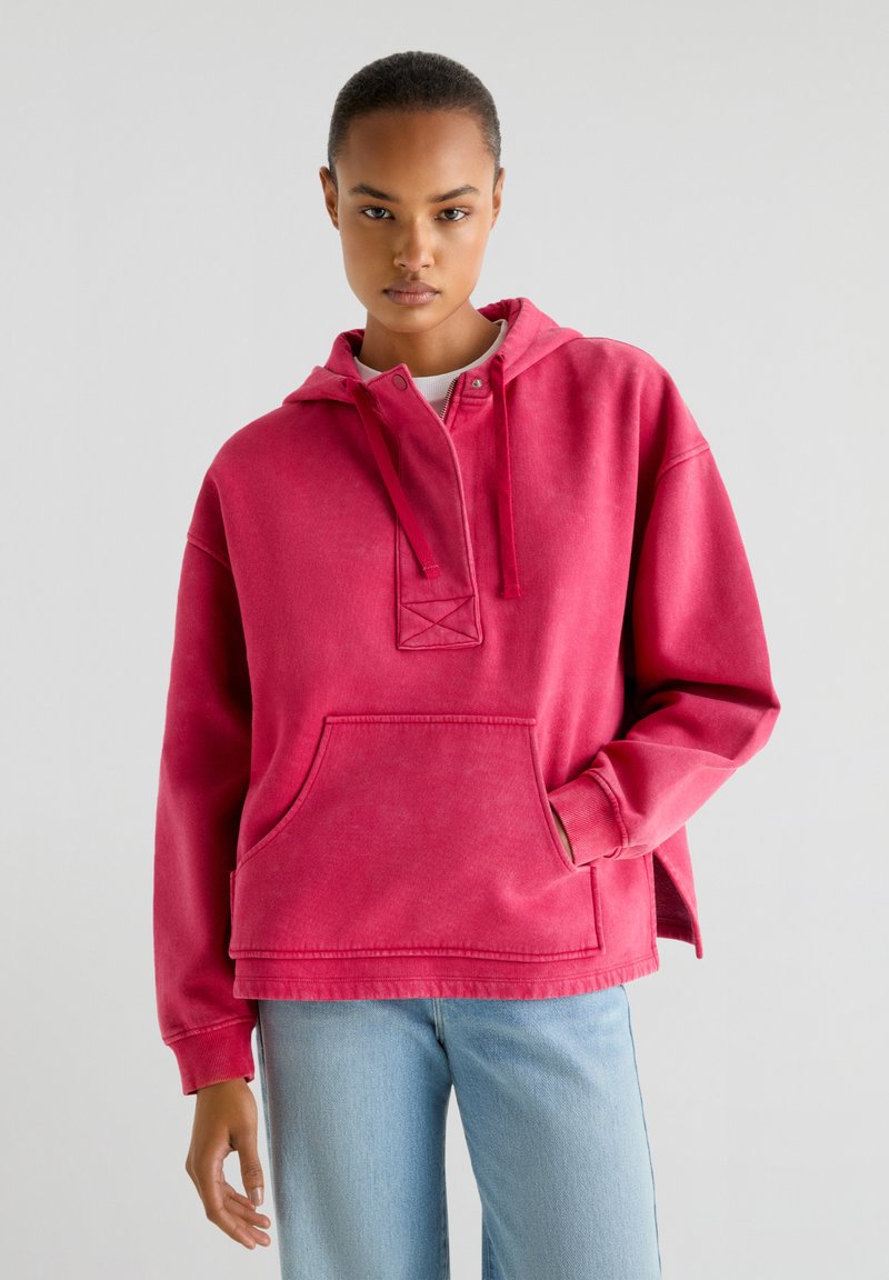 Haut à capuche rose avec une poche avant, un capuchon à cordon de serrage et une patte de boutonnage à moitié. Il a une coupe décontractée et un tissu doux et texturé.
