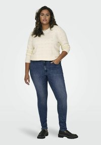 Light cream knit sweater with subtle texture, paired with dark blue skinny jeans and black slip-on shoes. Model stands with one hand on hip.