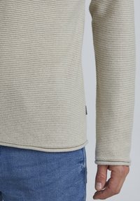 Hellbeige gestrickter Pullover mit strukturiertem, geripptem Muster. Hat lange Ärmel und einen abgerundeten Saum, kombiniert mit blauen Jeans.