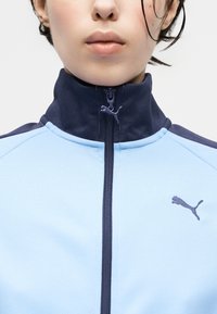 Κοντινό πλάνο ενός ατόμου που φοράει μπουφάν με φερμουάρ σε ανοιχτό μπλε και ναυτικό μπλε, με ψηλό γιακά και το λογότυπο Puma στο στήθος και στην τρύπα του φερμουάρ.