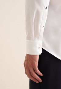 Manica di una camicia bianca con bottoni, caratterizzata da un polsino su misura con due chiusure a bottone e un dettaglio floreale ricamato vicino al polsino.