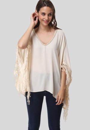 Túnica beige de corte amplio con escote en V, que presenta intrincados detalles de encaje en las mangas y el dobladillo. Textura suave, diseño informal.