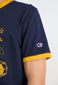 Marineblaues T-Shirt mit gelbem, geripptem Kragen und Ärmeln. Mit einem gestickten Logo auf dem linken Ärmel und einem gelben Grafikdruck. Aus einem Baumwollgemisch.