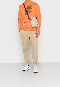 Man som bär en orange Nike-tröja, beige sweatpants, vita Nike-sneakers och en ljusgrå axelväska med brun rem mot en enkel vit bakgrund.