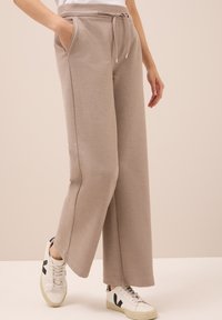 Beige wijde broek met een trekkoord in de taille en zijzakken, gedragen met witte sneakers met zwarte accenten.