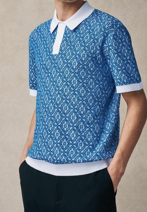 Homme portant un polo à manches courtes à motifs bleus avec un col et des poignets blancs, les mains dans les poches d'un pantalon noir, fond neutre.