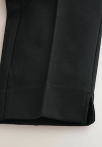 Nahaufnahme von schwarzem Stoff mit einer maßgeschneiderten Naht und einem kleinen seitlichen Schlitz am Saum vor einem neutralen Hintergrund.