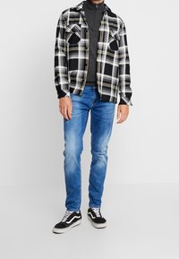 Schwarz-weiß kariertes Flanellhemd über einem dunkelgrauen Zip-up, kombiniert mit hellblauen Jeans und schwarzen Sneakern mit weißen Akzenten.