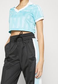 Kvinna som bär en ljusblå Adidas kortärmad sporttopp och svarta trackpants med ljusblå och vita sidostripes, stående med handen i fickan.