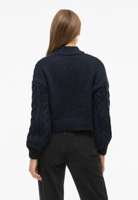 Granatowy sweter dzianinowy o grubym splocie, w krótkim kroju, z rękawami w plecionkę. Posiada wysoki dekolt i ściągacz przy dolnej krawędzi.