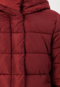Veste doudoune rouge avec une texture matelassée, une capuche et une fermeture éclair à l'avant. Le matériau semble lisse et légèrement brillant avec des sections rembourrées.