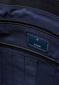 Interno di una borsa in tessuto blu navy con dettagli in pelle nera strutturata e un'etichetta blu del marchio etichettata "JOOP!" con accenti metallici.