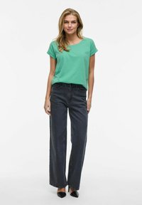 Grön kortärmad t-shirt i kombination med högmidjade mörkgrå vida jeans och svarta spetsiga klackskor. Tyget verkar slätt och avslappnat.