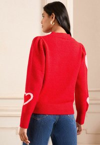 Jersey de punto rojo con mangas abullonadas, que presenta patrones de corazones blancos en los puños y un dobladillo acanalado. Tejido grueso y texturizado.