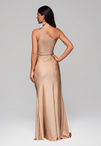 Un vestido de noche beige de un solo hombro con un corsé ajustado, falda fluida y detalle decorativo en el hombro. Tela suave y ligeramente brillante.