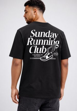 Mann mit dem Rücken zur Kamera trägt ein schwarzes T-Shirt mit weißem Text „Sunday Running Club“ und einer geflügelten Schuh-Grafik auf dem Rücken.