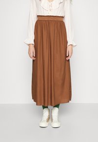 Donna con una gonna midi marrone a vita alta, blusa bianca con motivo e maniche a sbuffo, calzini verdi e sneakers bianche alte.