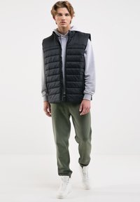 Černá prošívaná vesta s kapucí, kombinovaná se šedou mikinou a joggerkami v olivově zelené barvě. Bílé tenisky dokončují outfit.