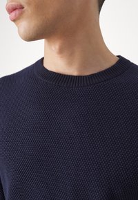 Marinblå texturerad stickad tröja med ribbad rundhals, som har ett upphöjt mönster och en slät yta.