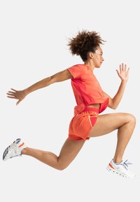 Conjunto deportivo rojo que incluye una camiseta de manga corta y pantalones cortos. Zapatillas de running blancas con acentos naranjas. El modelo está en una pose dinámica de carrera.