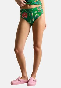 Žena v zelených květovaných vysokých bikinách a růžových Birkenstock klopkách, stojící na pozadí s bílou plochou.