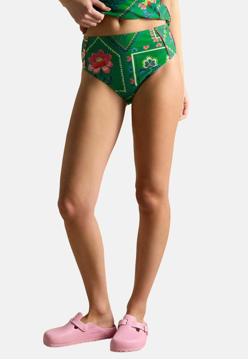 Žena v zelených květovaných vysokých bikinách a růžových Birkenstock klopkách, stojící na pozadí s bílou plochou.