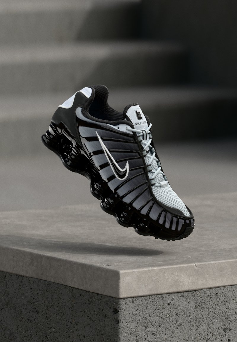 Chaussure de course Nike noir et blanc suspendue au-dessus d'une surface en béton avec un arrière-plan de pierres en escalier.