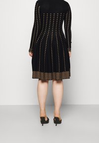 Robe noire à manches longues avec un motif de losanges dorés et des accents texturés, s'évasant à l'ourlet. Portée avec des sandales noires à talons.