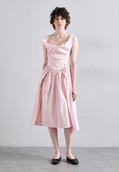 Robe en coton rose avec un décolleté carré, taille froncée et jupe évasée. Présente des bretelles larges et un petit détail de logo. Chaussures noires plates.