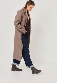 Long, brown wool coat with a collar, worn over a dark vest and blue denim jeans. Featuring gray ankle boots with a smooth finish.