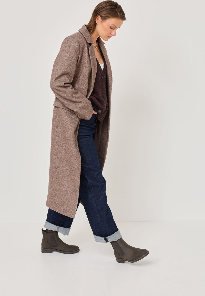 Long, brown wool coat with a collar, worn over a dark vest and blue denim jeans. Featuring gray ankle boots with a smooth finish.