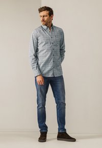 Hellblaues Hemd mit kleinem, sich wiederholendem Muster. Lange Ärmel, Standardkragen. Kombiniert mit dunklen Jeans und braunen Schuhen.