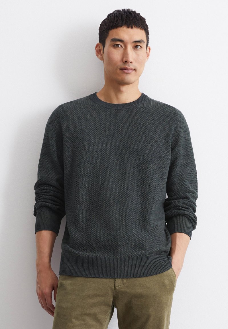 Sweater in dark green knit with a textured pattern, crew neck, and ribbed cuffs. Paired with khaki pants.