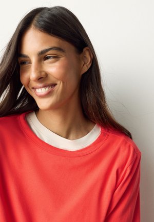 Mladá žena s dlhými tmavými vlasmi sa usmieva a pozerá sa bokom, oblečená v červenej košeli nad bielym tričkom s okrúhlym výstrihom, na jednoduchej svetlej pozadí.