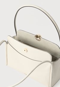 Kremasta usnjena torbica z zaponko v zlati barvi, žepom z zadrgo notranjosti in okroglo naramničo na bele podlagi.