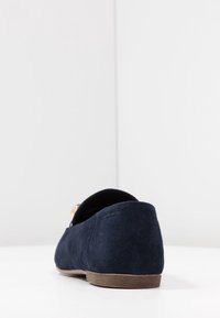 Zapato plano de ante azul marino con una silueta redondeada, diseño minimalista, textura suave y una pequeña suela de goma marrón.