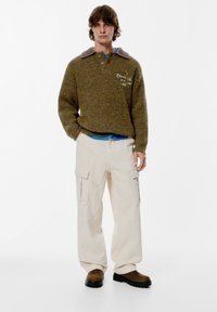 Ruskea teksturoitu neulepaita, jossa on violetti kaulus, napituslista ja kirjailtuja yksityiskohtia, yhdistettynä kermaisiin cargo-housuihin ja ruskeisiin saappaisiin.