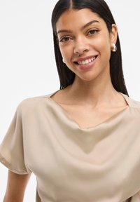 Blouse en soie beige avec un décolleté large et peu profond et des manches courtes. Texture lisse avec un design drapé, mettant en avant la douceur et le mouvement.