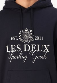 Hoodie blu navy con testo bianco "LES DEUX Sporting Goods" e logo a corona. Realizzato in tessuto morbido, ha una tasca frontale e cappuccio regolabile.
