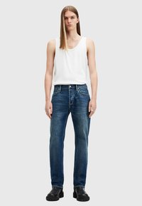Jeans de denim azul con corte recto, cinco bolsillos y una clásica bragueta de botones. Llevados con una camiseta sin mangas blanca y botas negras de tobillo.