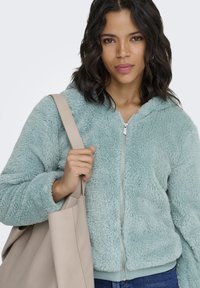 Flauschiger, petrolfarbener Kapuzenpullover mit Reißverschluss, Kapuze, geripptem Saum und Bündchen. Der Träger hält eine hellbeige Tragetasche, die die Textur der Jacke zeigt.