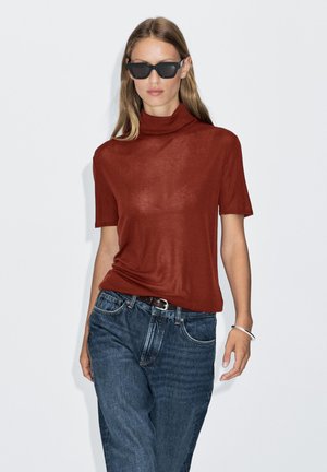 Top a collo alto a maniche corte color ruggine abbinato a jeans denim scuro, con vestibilità rilassata e una leggera lucentezza. Accessoriato con occhiali da sole neri.