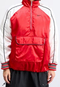 Person klädd i en röd och vit sportig zip-up-jacka med framficka, svarta byxor och randiga muddar på ärmarna.