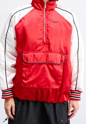 Personne portant une veste zippée sportive rouge et blanche avec une poche avant, un pantalon noir et des poignets rayés sur les manches.